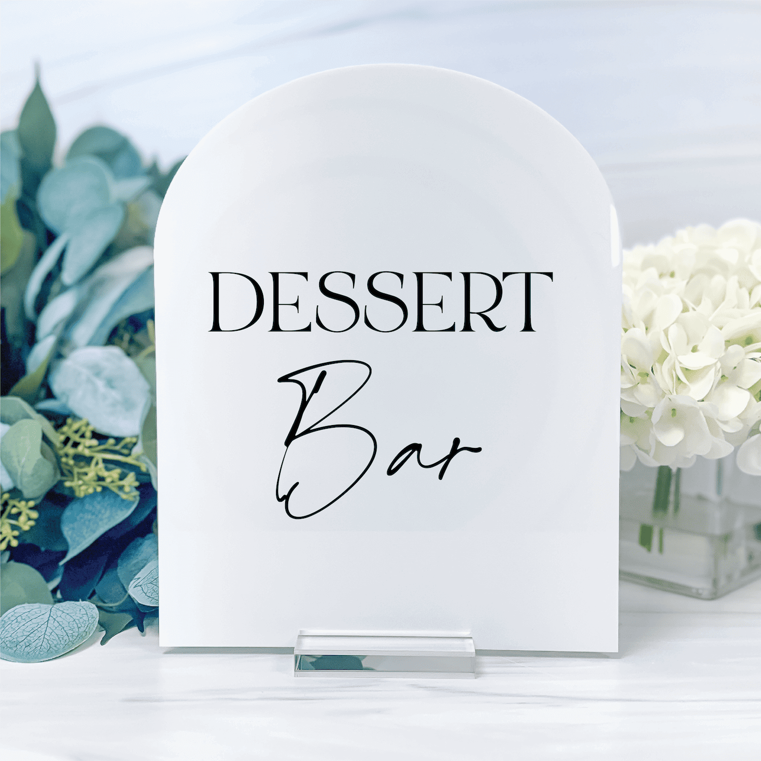 White arch-shaped acrylic sign reading "DESSERT Bar" in black script text, displayed with flowers and greenery.