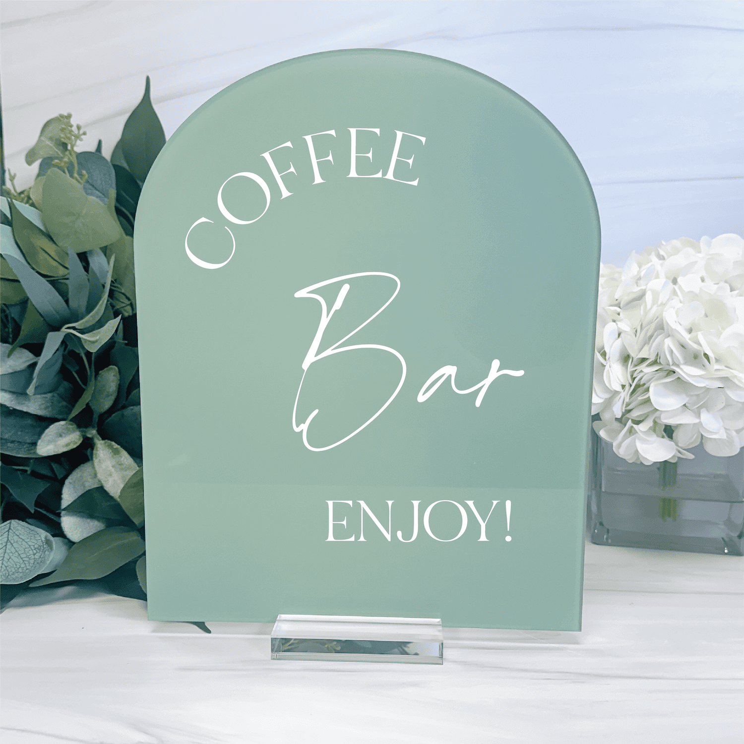 Sage green acrylic table sign with white text reading "Coffee Bar Enjoy" and decorative eucalyptus beside it
