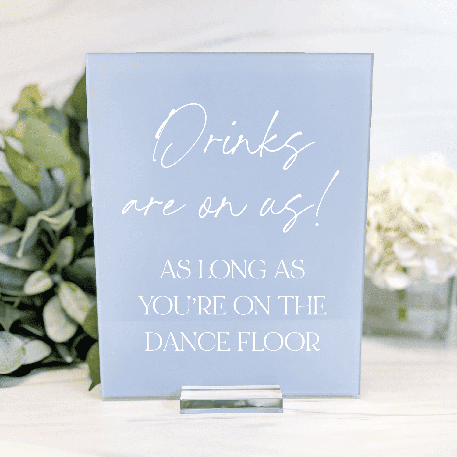 Light blue acrylic sign with white script and block text reading "Drinks are on us! As long as you're on the dance floor" next to greenery and flowers.
