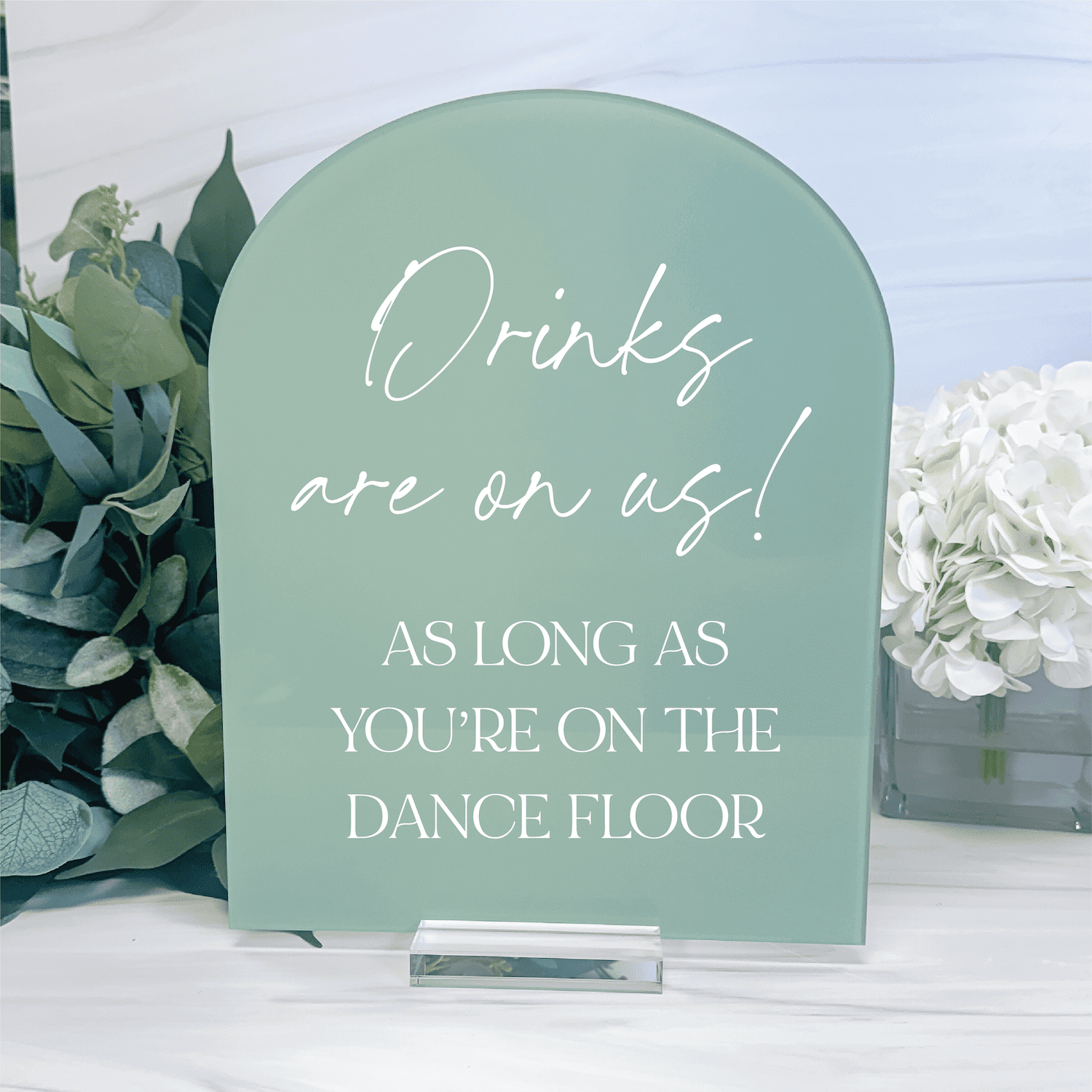 Sage green acrylic arch sign with white script and uppercase text reading "Drinks are on us! AS LONG AS YOU'RE ON THE DANCE FLOOR" surrounded by greenery and white flowers.