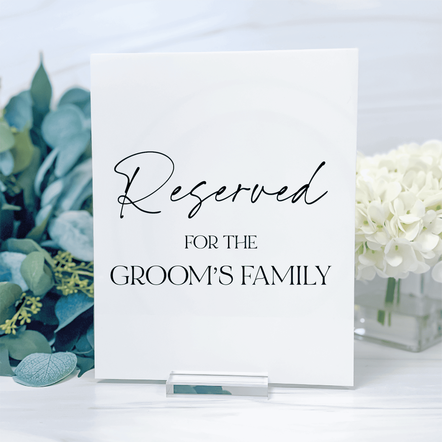 White acrylic table sign with "Reserved for the Groom's Family" text, displayed among white flowers and greenery