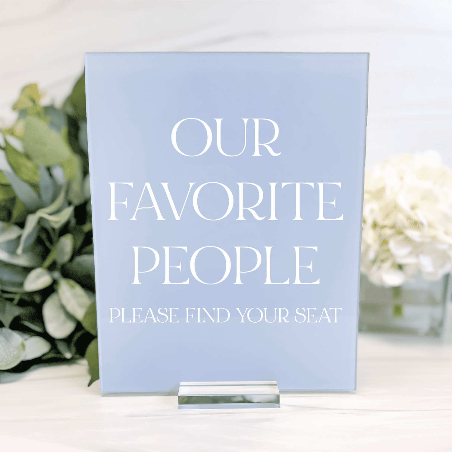 Light blue acrylic seating sign with white text reading "Our Favorite People Please Find Your Seat" surrounded by greenery and white flowers.