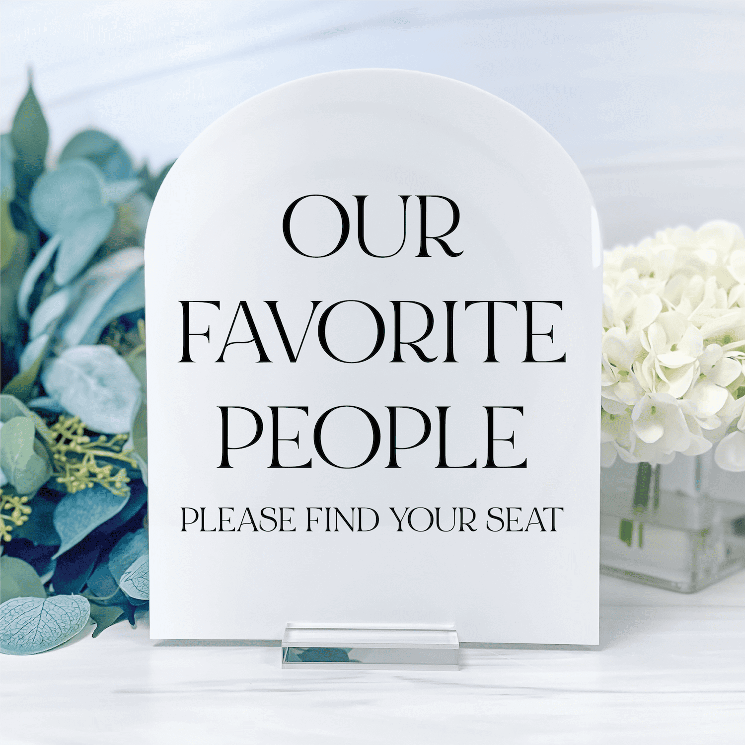 White acrylic arch-shaped seating sign reading "Our Favorite People Please Find Your Seat" with eucalyptus and white flowers in background