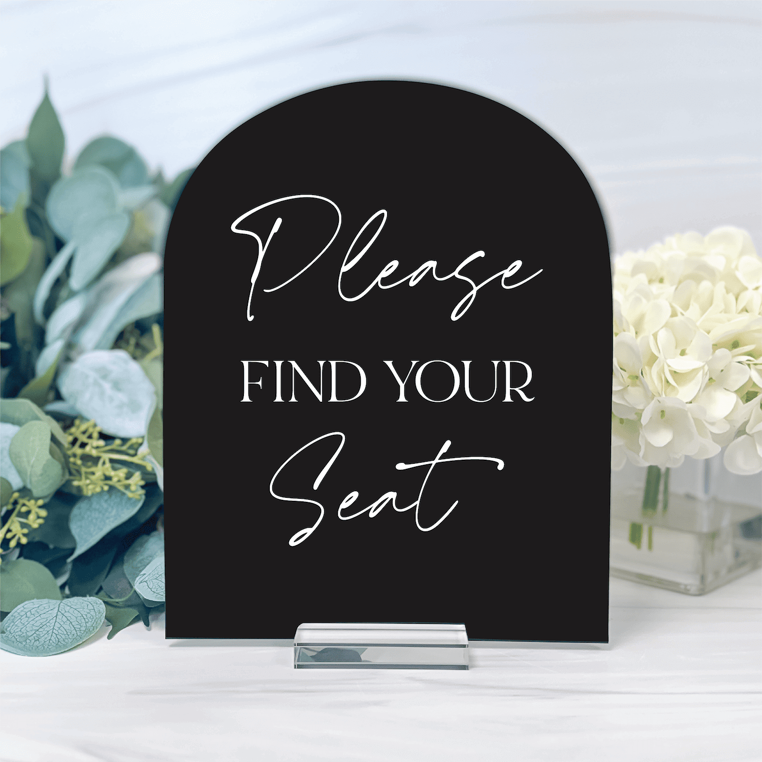 Black arched acrylic sign with "Please FIND YOUR Seat" in white script and sans-serif lettering, displayed on a table with white flowers and greenery.