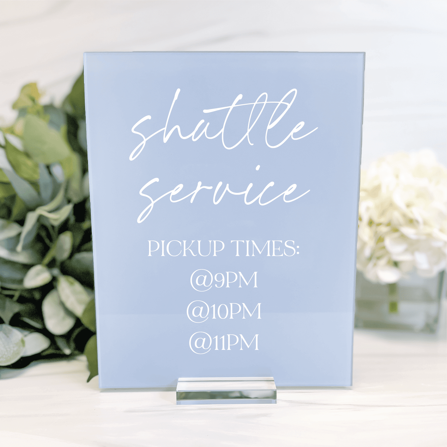 Light blue acrylic sign with white cursive text reading "shuttle service" and pickup times listed below, displayed with flowers and greenery.