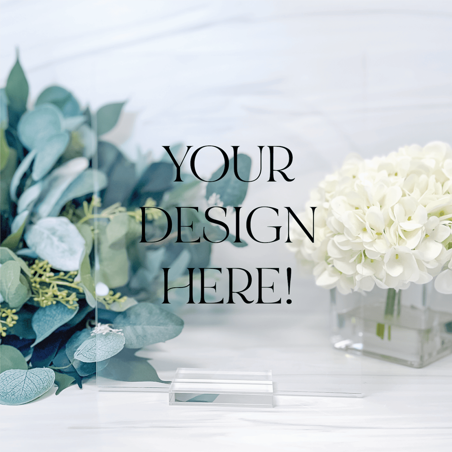 Clear acrylic sign with "YOUR DESIGN HERE!" text displayed on white surface among eucalyptus and white hydrangea flowers