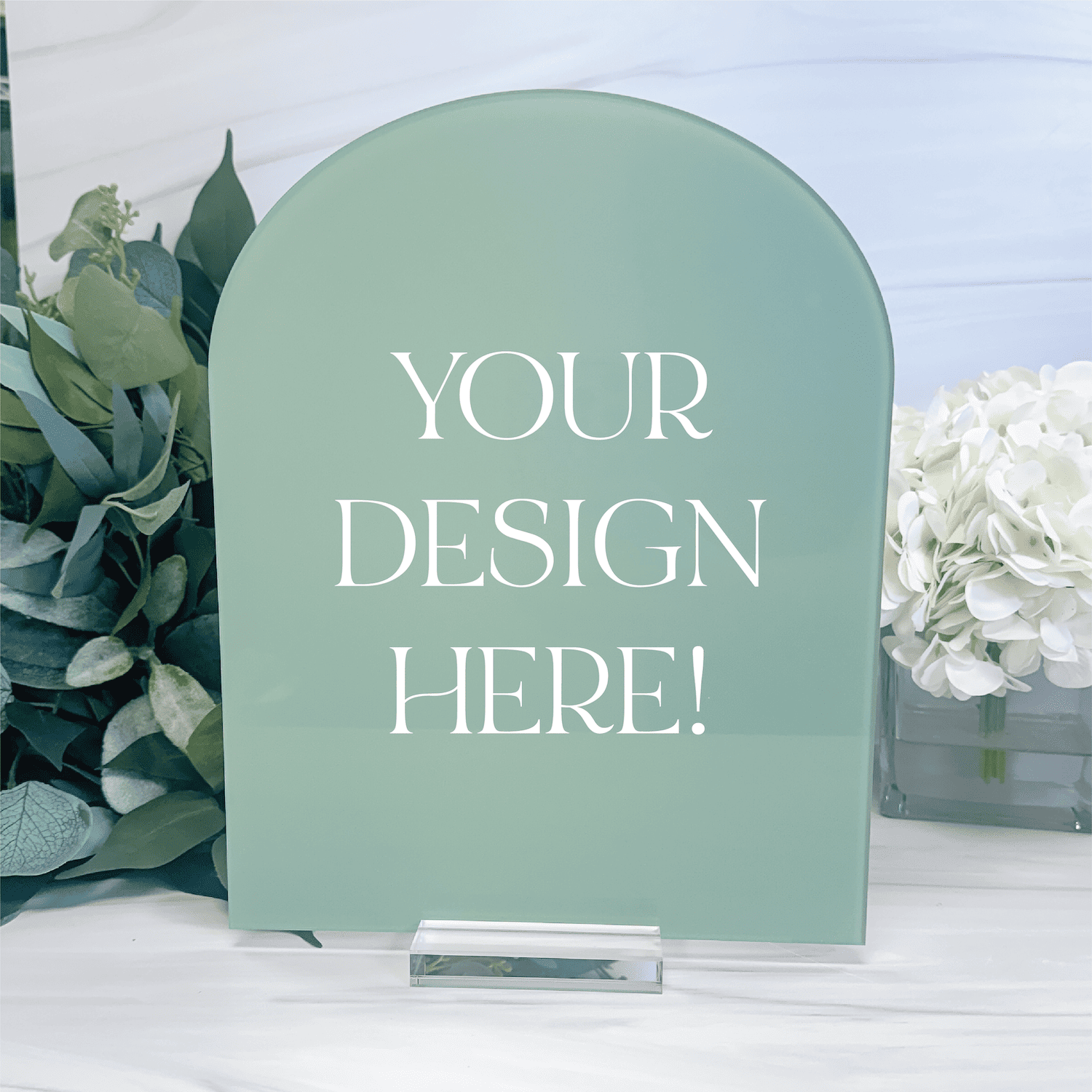 Sage green arched acrylic sign with "YOUR DESIGN HERE!" text, displayed on white surface with greenery and white flowers.