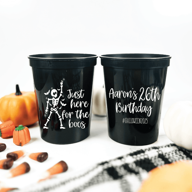 Two black stadium cups with white text, one reading "Just here for the boos" with skeleton design, the other personalized "Aaron's 26th Birthday #HalloweenBirthday," surrounded by Halloween decorations and pumpkins.