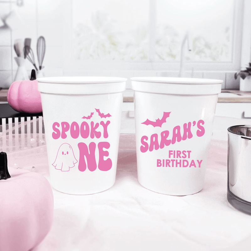 White stadium cups with pink text, one reading "SPOOKY ONE" with ghost design, other reading "SARAH'S FIRST BIRTHDAY" with bat graphic