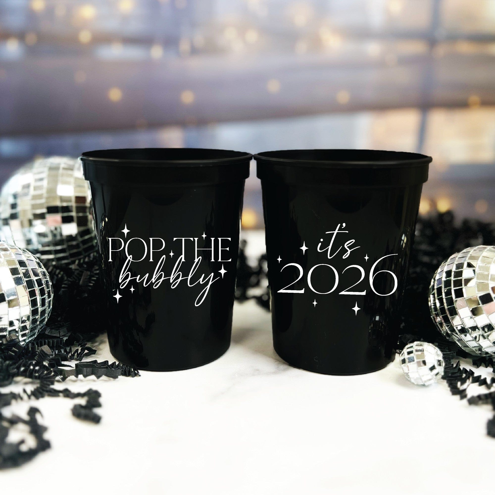 Two black plastic party cups with white festive designs, arranged with disco balls and black crinkle paper, suitable for a celebratory event.
