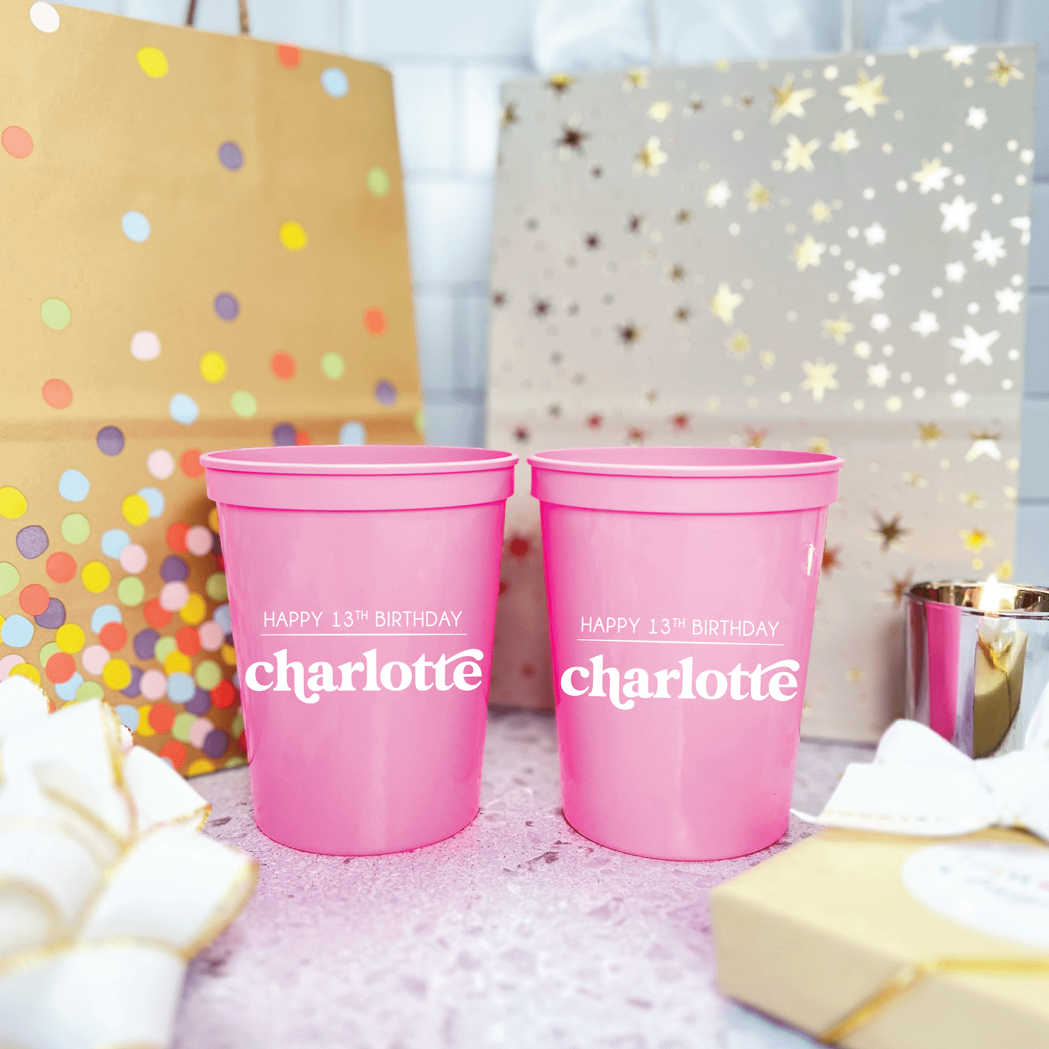 Bright pink stadium cups personalized with "Charlotte" and "Happy 13th Birthday" text, displayed with colorful polka dot and star backdrop decorations.