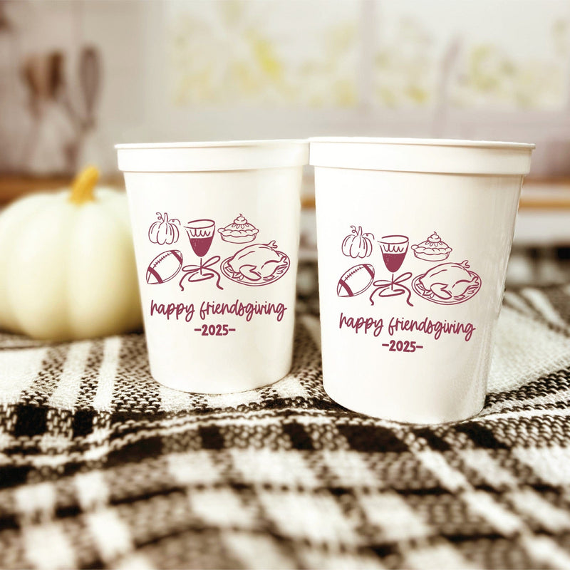 Two white plastic stadium cups display a maroon autumn-themed graphic featuring seasonal food, drink, and sports elements, perfect for fall gatherings.