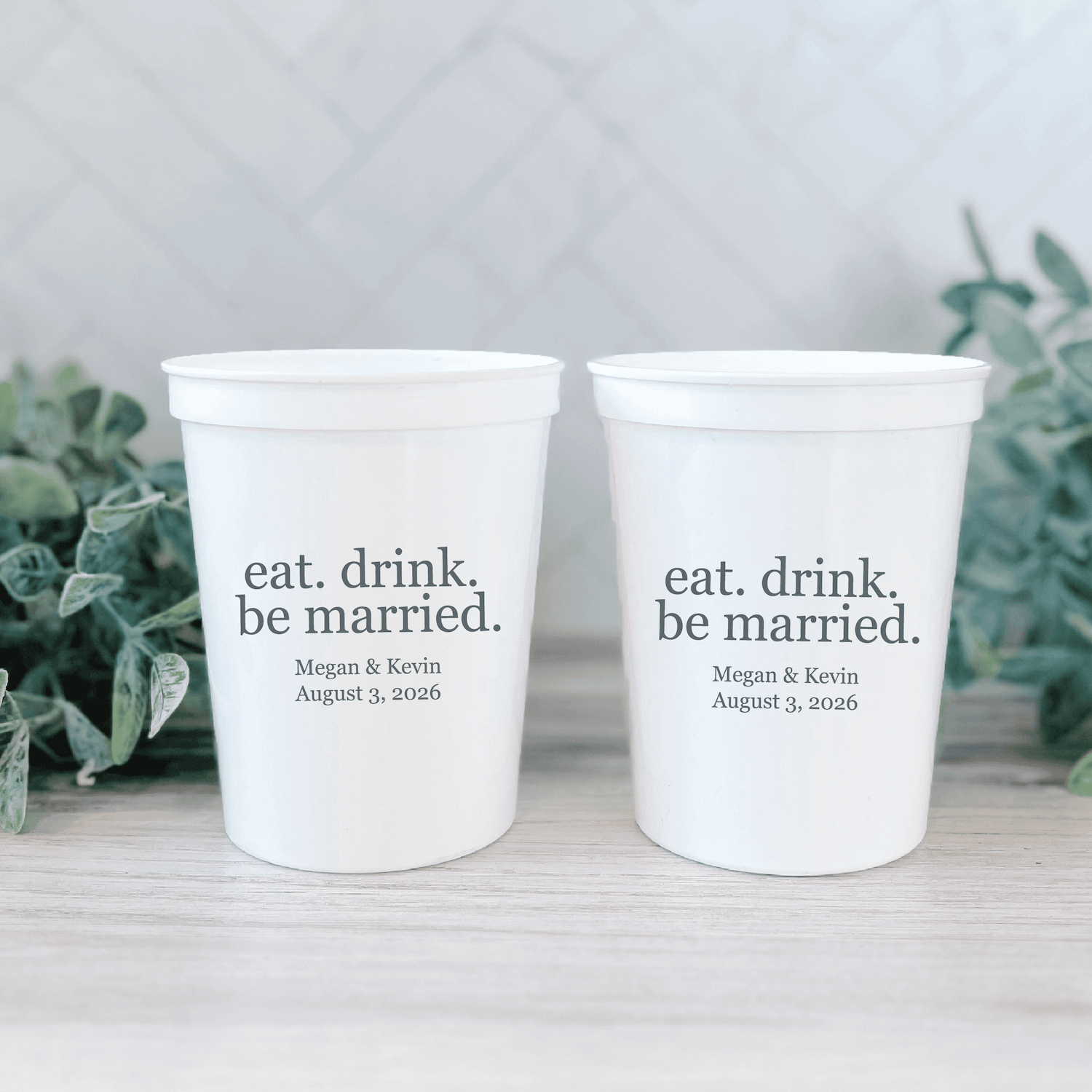 Two white stadium cups with "eat. drink. be married." text and personalized names and wedding date printed in black.