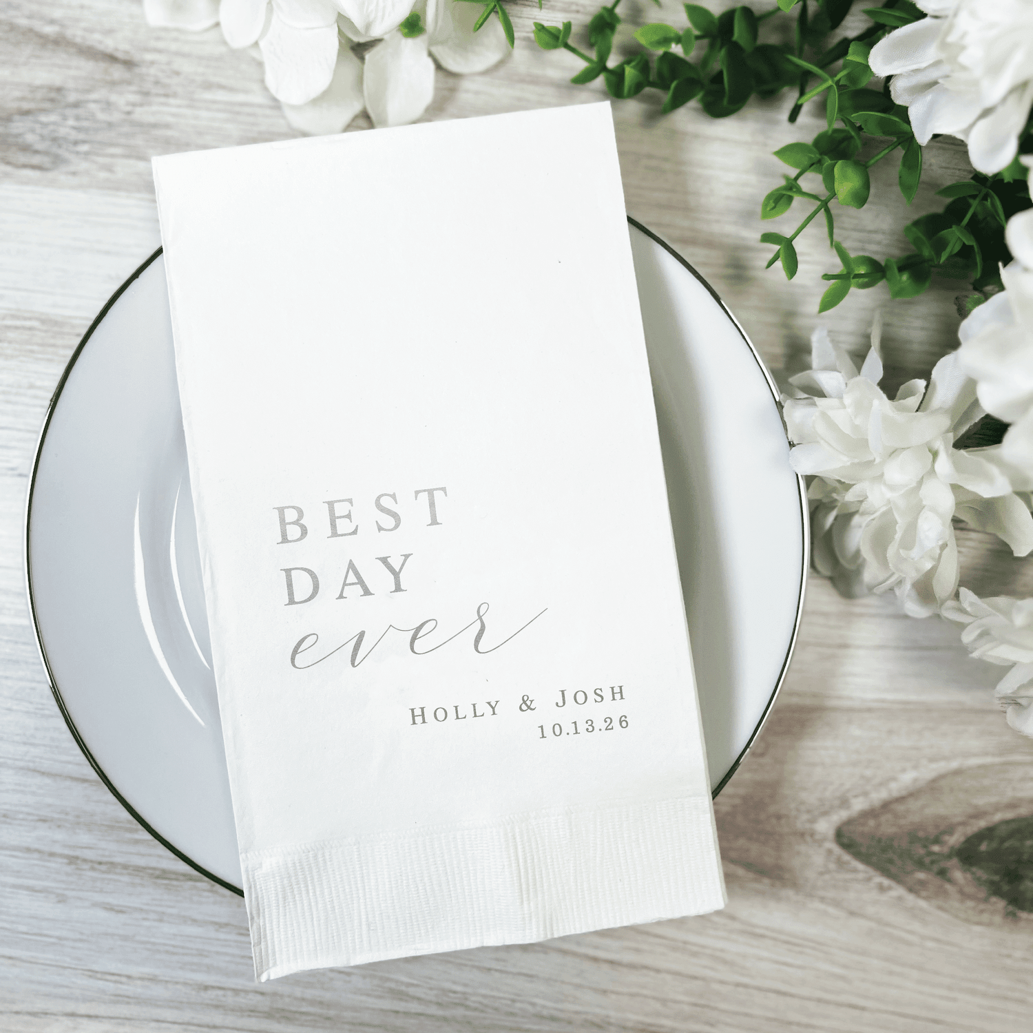 White napkin with "Best Day Ever" text and couple names Holly and Josh with date, placed on gray plate with white flowers
