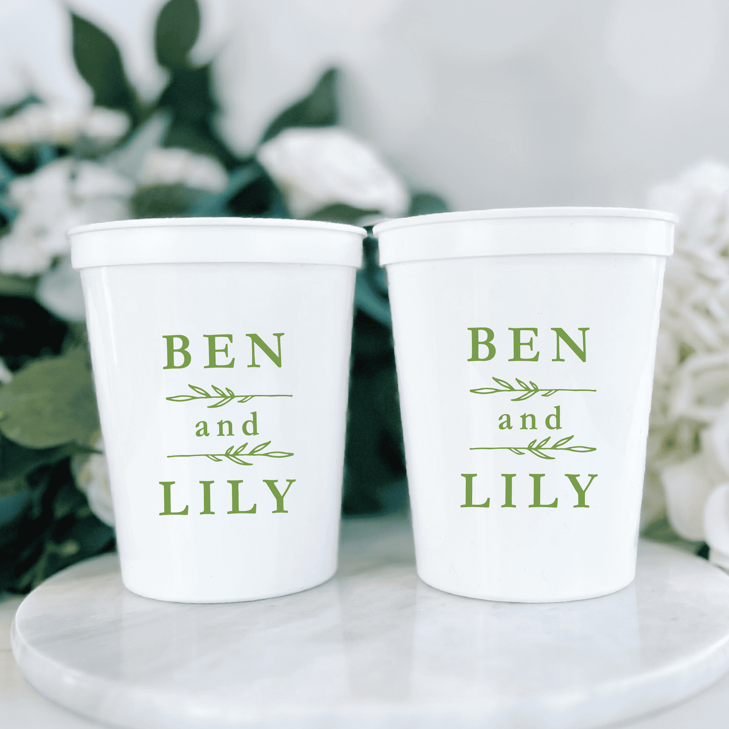 Two white stadium cups with green text reading "BEN and LILY" displayed on a white marble surface with greenery in background.