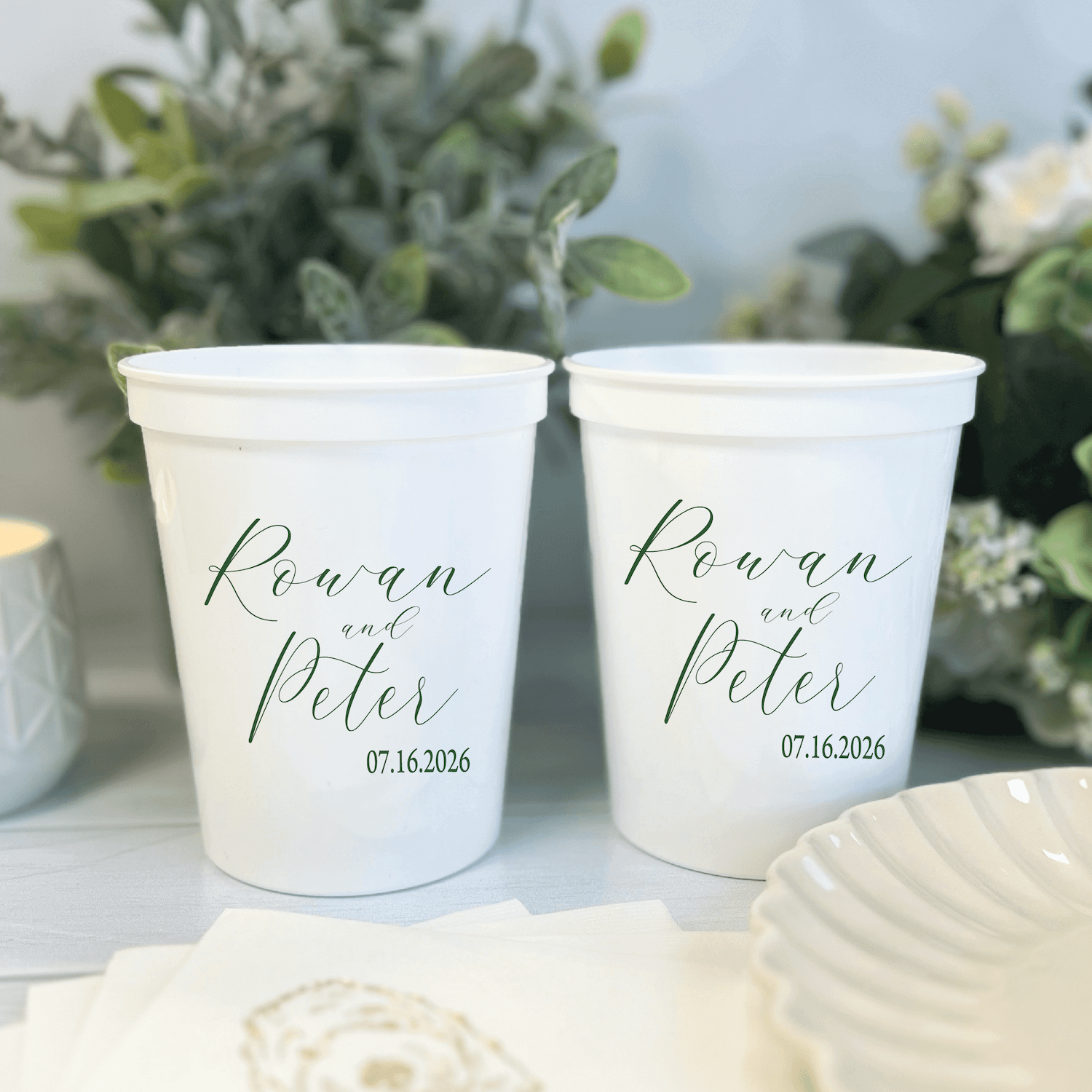 Two white stadium cups personalized with "Rowan and Peter" in script and the date 07.16.2026, displayed with greenery and white flowers.