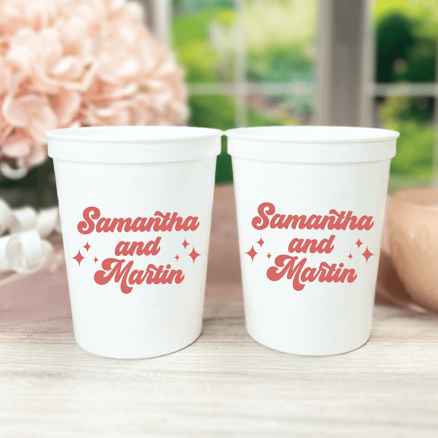 Two white stadium cups with red retro script text reading "Samantha and Marin" with decorative sparkle designs, displayed outdoors with flowers.