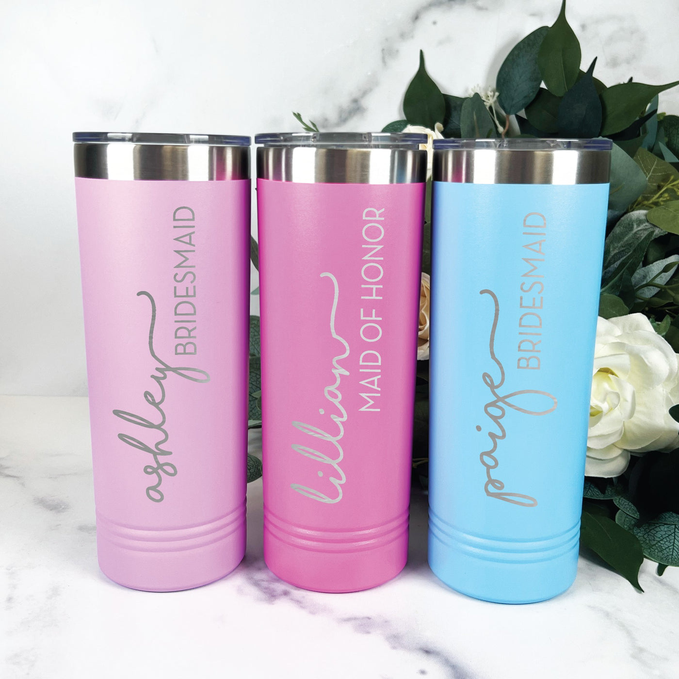 Three personalized stainless steel tumblers in varying shades of pink and blue, featuring a clear lid and metal rim, perfect as bridal party gifts for a wedding.