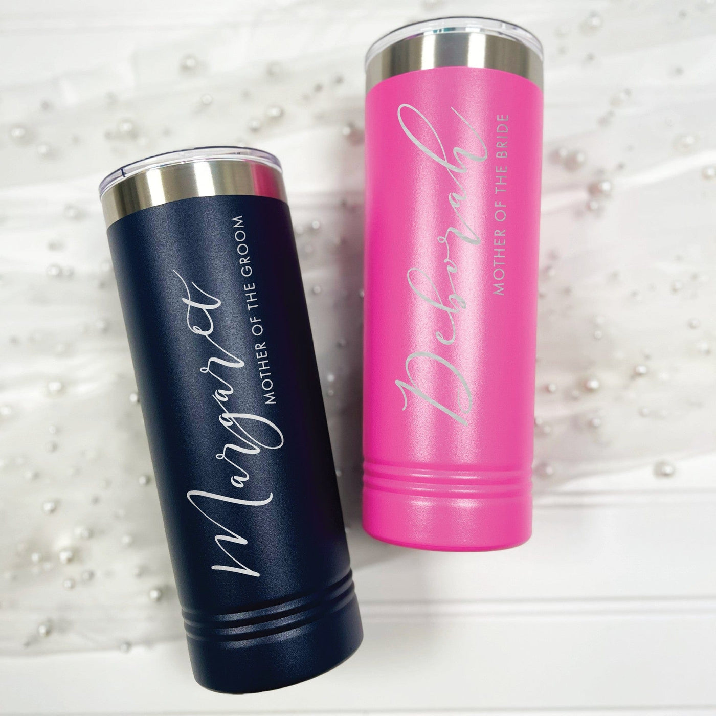 Two personalized skinny insulated tumblers, one navy blue and one hot pink, made of metal with clear lids, designed as wedding party gifts and displayed on a white surface with pearl accents.