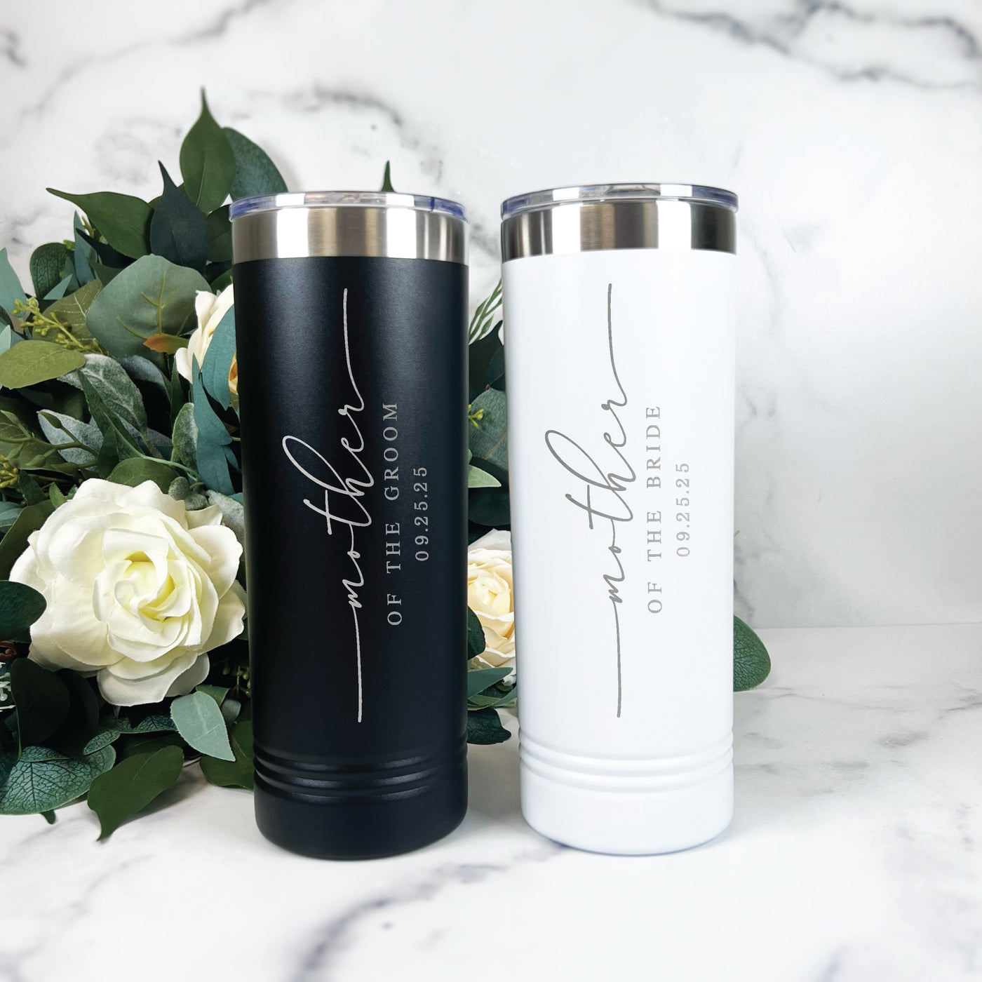 A pair of personalized insulated stainless steel tumblers, one black and one white, each with a silver rim and clear sliding lid, designed as thoughtful gifts for a wedding event.
