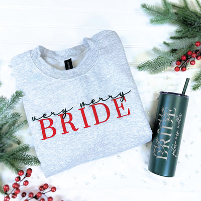 A light gray crewneck sweatshirt and a dark green insulated tumbler, both personalized for a bride with a 'very merry BRIDE' design, displayed with festive holiday greenery and berries.