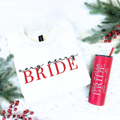 A cozy light grey crewneck sweatshirt and a matching red insulated tumbler with a straw, both customized with a holiday bridal theme, presented amidst winter greenery and red berries for a festive touch.
