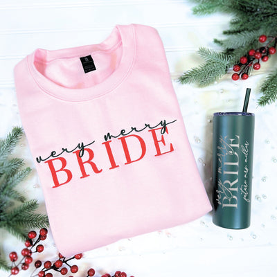 A pink crewneck sweatshirt and a dark green skinny tumbler, both personalized with festive bridal-themed text, for a unique wedding or engagement gift.