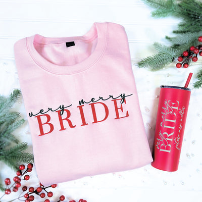 A light pink fleece crewneck sweatshirt with "very merry BRIDE" embroidery in red and black, paired with a red stainless steel tumbler featuring similar bridal-themed text, presented as a festive holiday gift set.