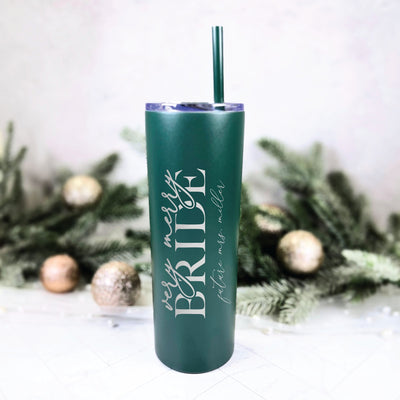 A dark green insulated tumbler with a matching straw features personalized laser-engraved bridal themed text, set against a festive background of evergreen branches and gold holiday ornaments, perfect for a winter wedding or bridal celebration.