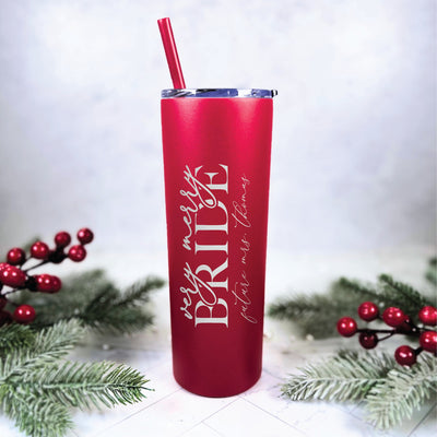 A red insulated metal tumbler with a clear lid and matching straw, featuring personalized white text, is styled with frosted pine branches and red berries for a festive bridal theme.