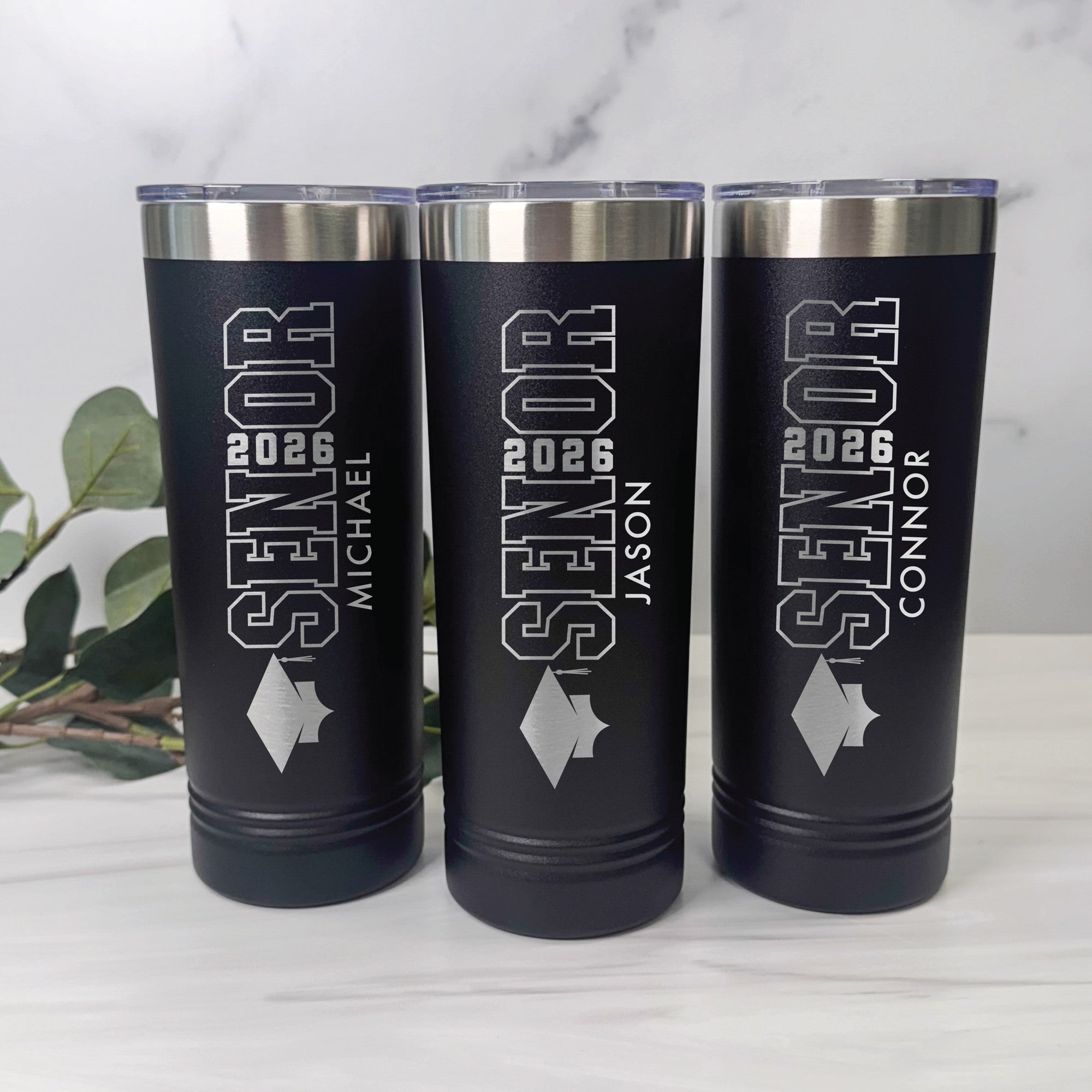 A set of three personalized black insulated tumblers with stainless steel rims and clear lids, engraved with a graduation theme for a graduation event.