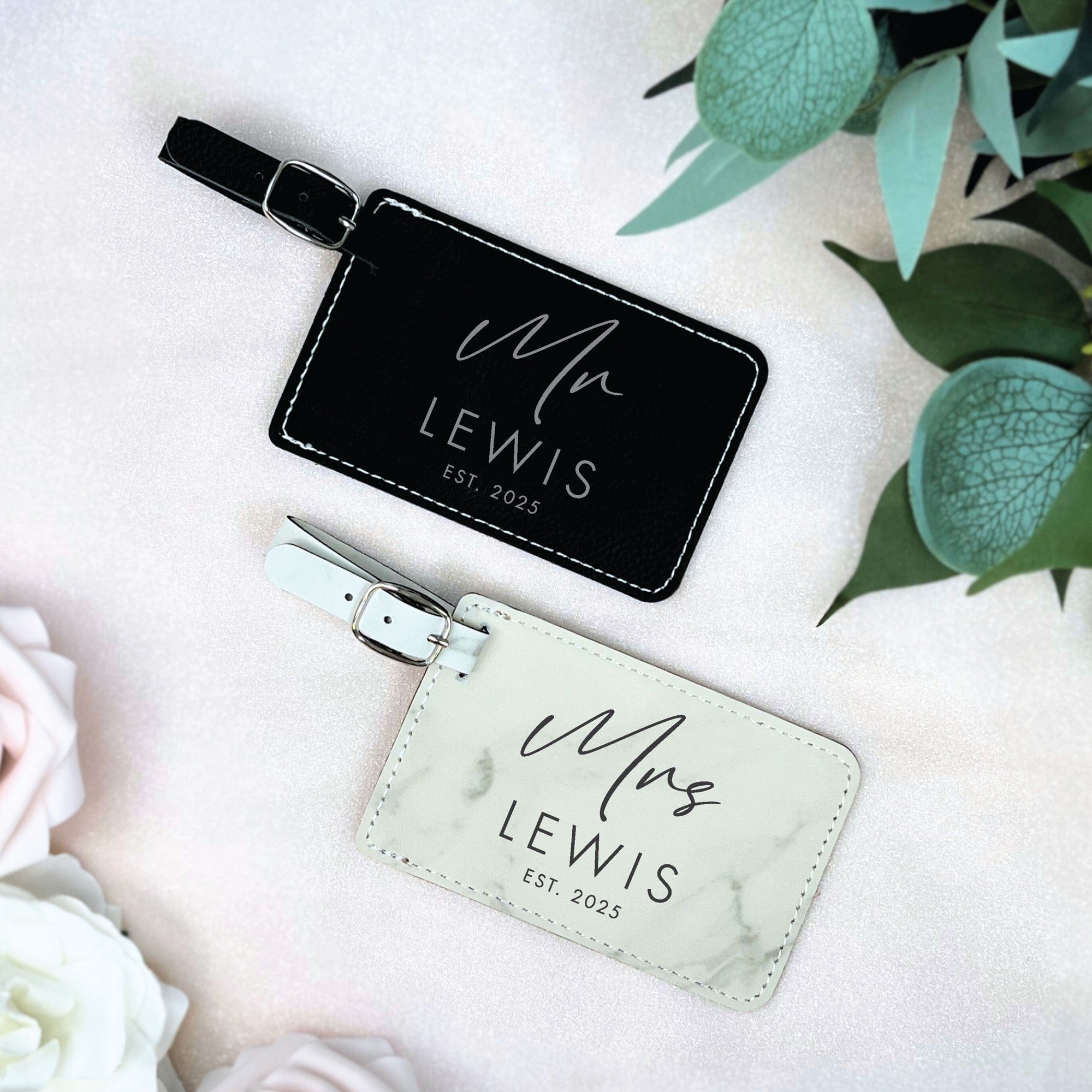 Two rectangular faux leather luggage tags, one black and one white marble pattern, with white stitching and metal buckles, designed as personalized travel accessories for a wedding or honeymoon.