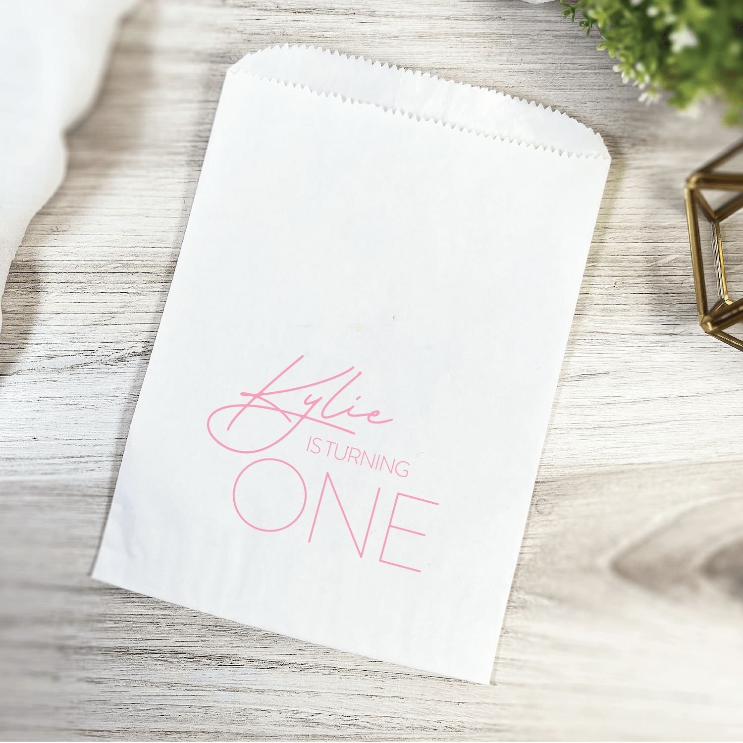 White treat bag with pink "She's Turning ONE" script text design on wood surface