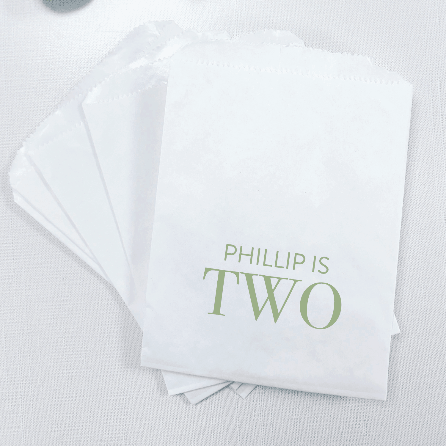 Stack of white treat bags with "Phillip is Two" printed in sage green text