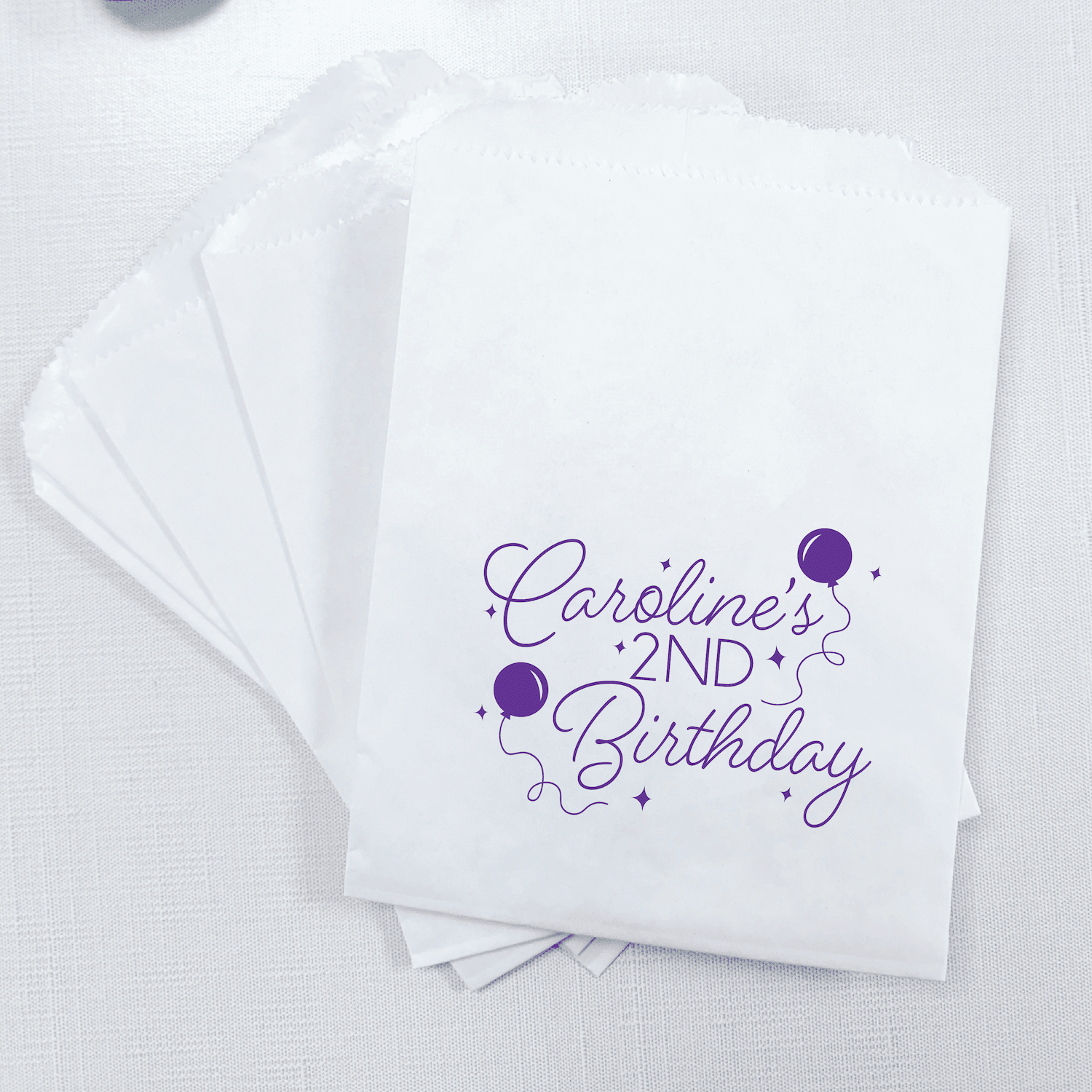 White treat bags with purple script text reading "Caroline's 2nd Birthday" and purple balloon designs