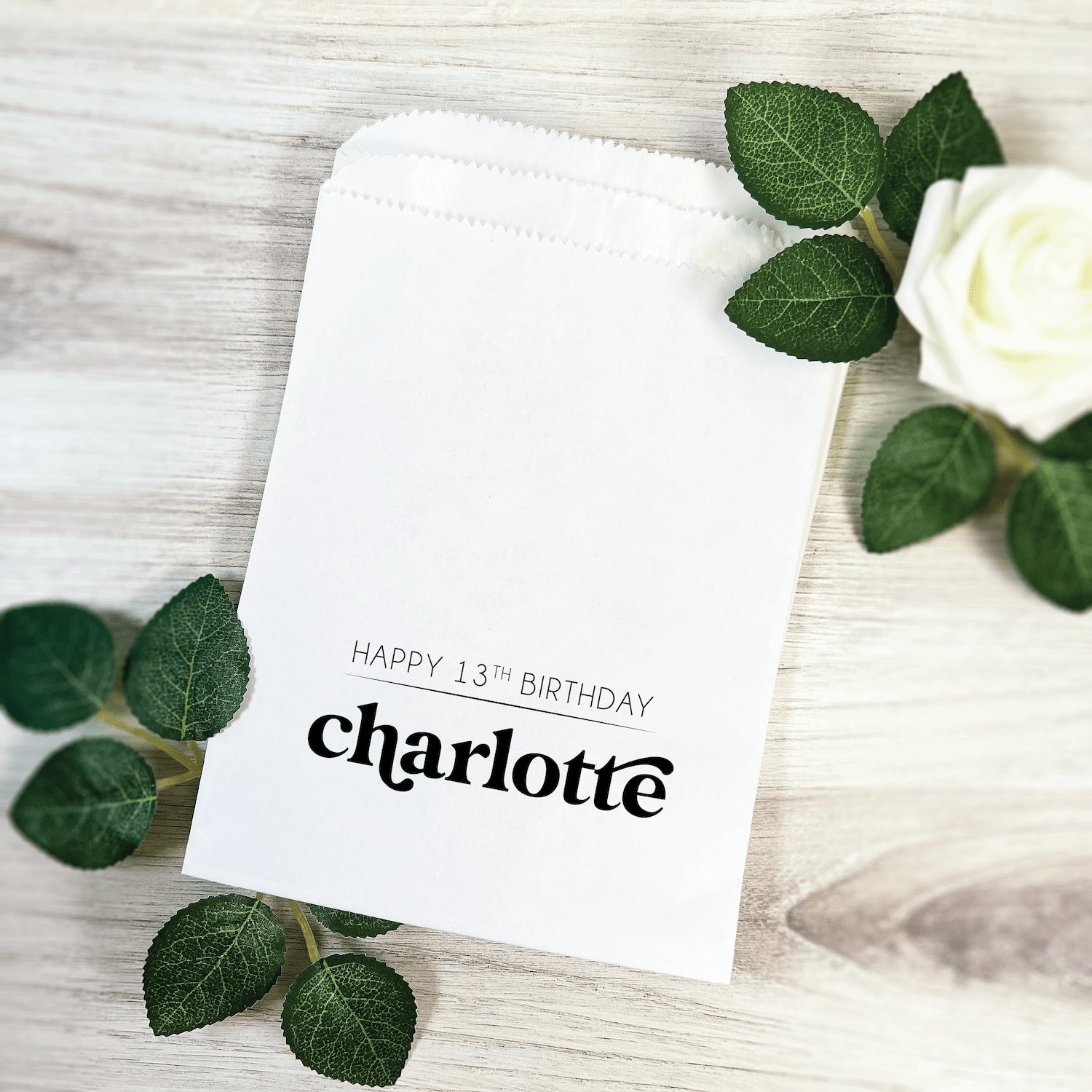 White treat bag with "Happy 13th Birthday Charlotte" personalized text, surrounded by green leaves and white flower on wooden surface