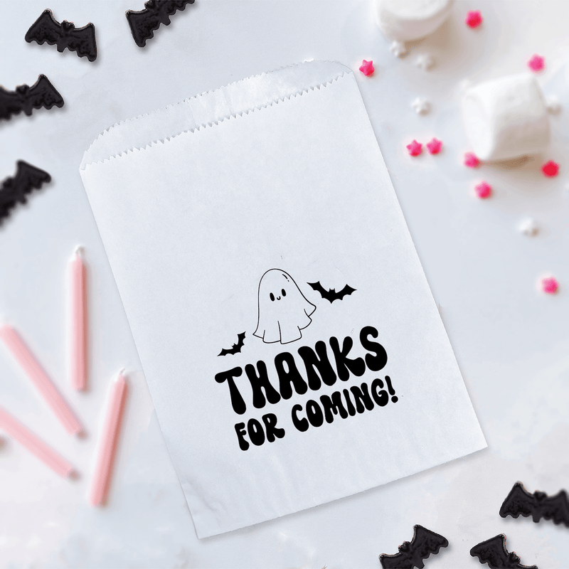 White treat bag with ghost and bats design reading "Thanks for Coming!" surrounded by Halloween decorations and party supplies.