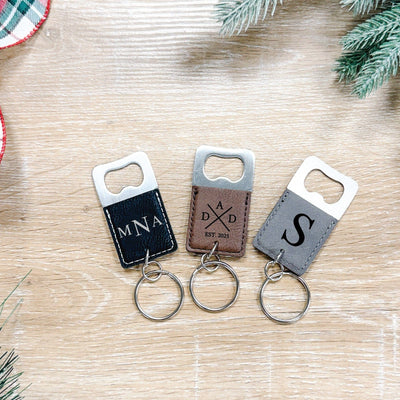 Three personalized bottle opener keychains featuring metal hardware and faux leather sleeves in black, brown, and grey, each with a custom engraved design and a silver keyring, perfect for event favors or gifts.