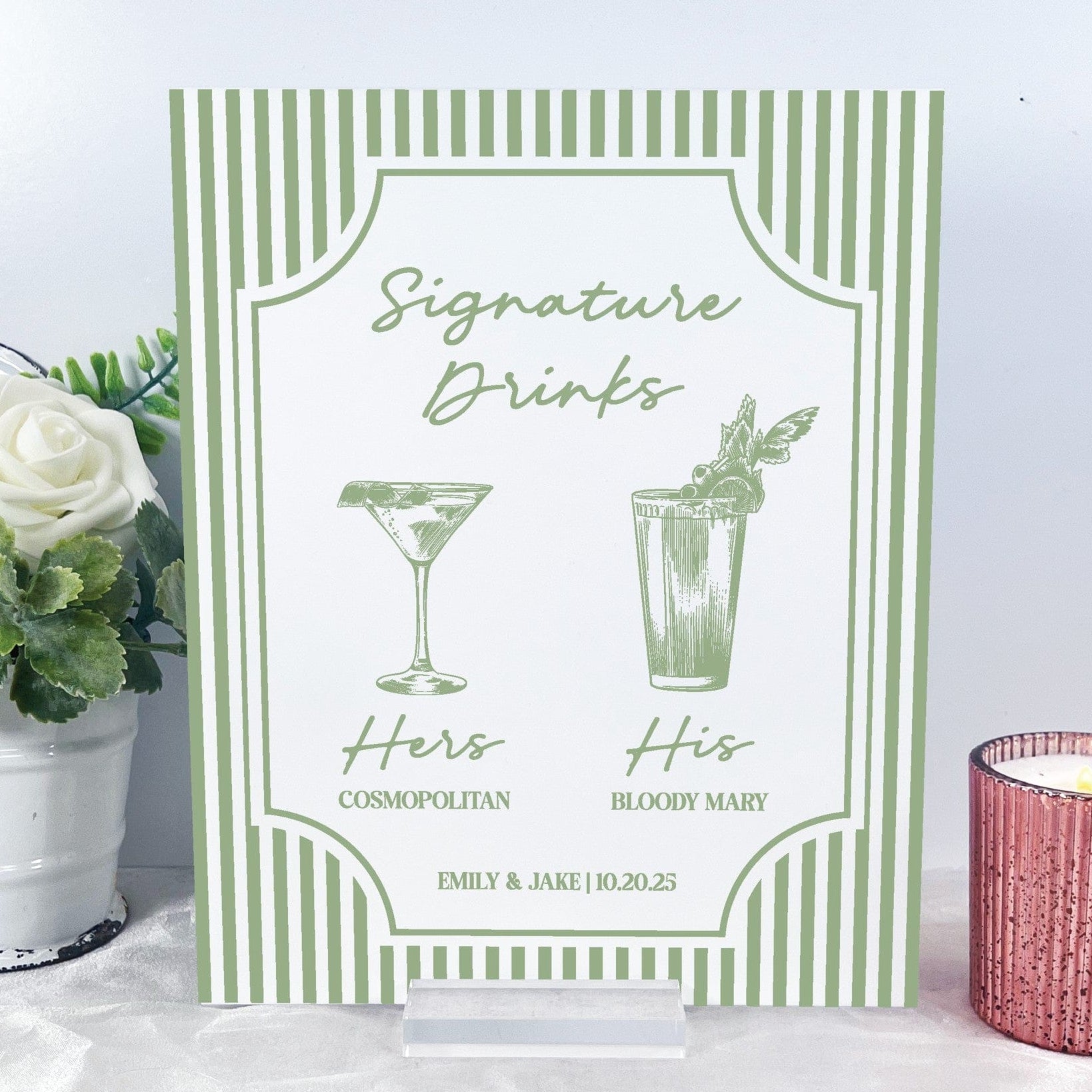 A personalized wedding signature drinks sign on a clear acrylic stand, featuring a white background with a green vertical stripe border and green script text for two signature cocktails with corresponding drink illustrations, perfect for an event bar.