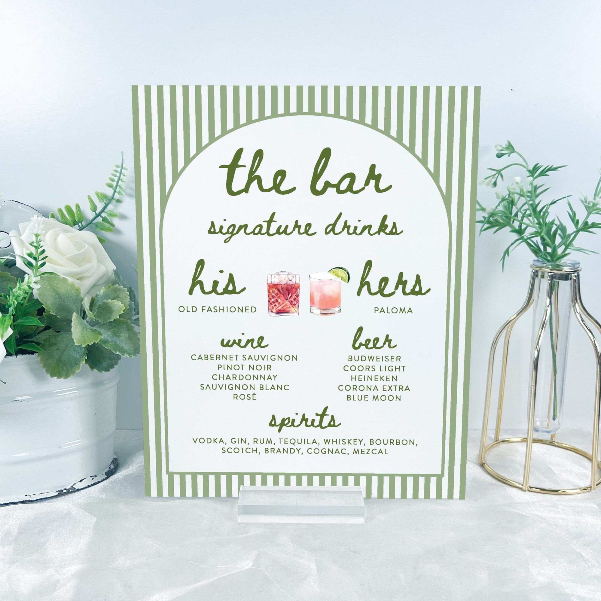 A rectangular acrylic bar menu sign featuring a sage green striped border and arched white center, sitting on a clear acrylic stand, displaying drink options for a wedding or event.