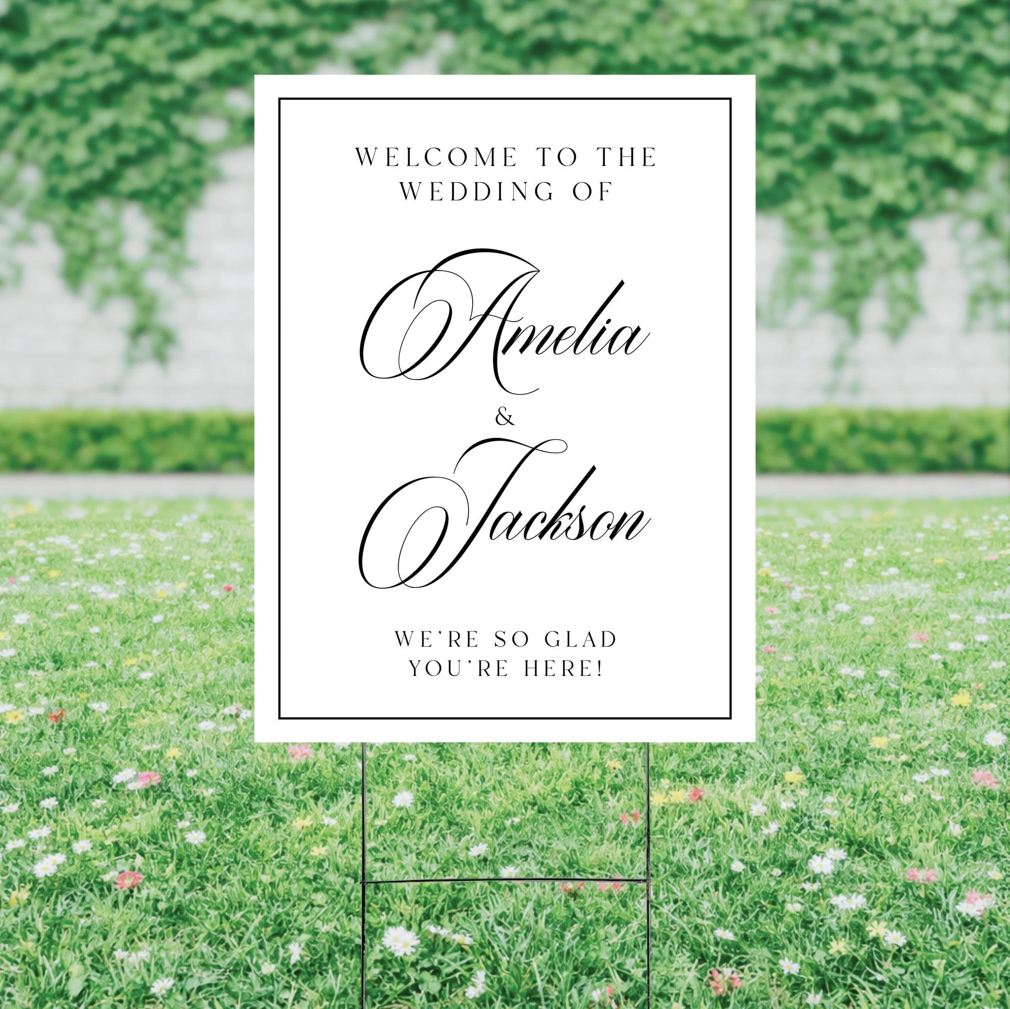 A white wedding welcome yard sign featuring elegant black script text, supported by a black metal H-stake, set amidst a green lawn with wildflowers.